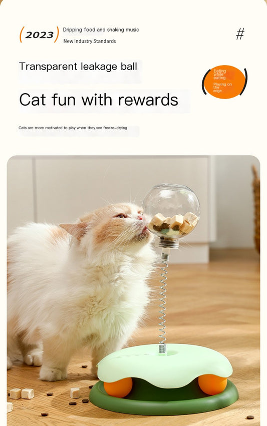 White cat playing with transparent treat-dispensing ball on spring and green base, with kibble scattered on floor