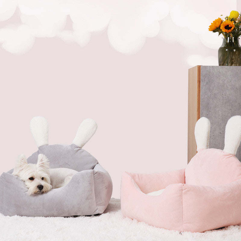 Two pet beds, one gray and one pink, with a small white dog lying on the gray bed in a room with a pink wall and a vase of flowers.